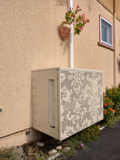 Cache climatiseur extérieur perforé beige installé sur une façade de maison avec fleurs et végétation